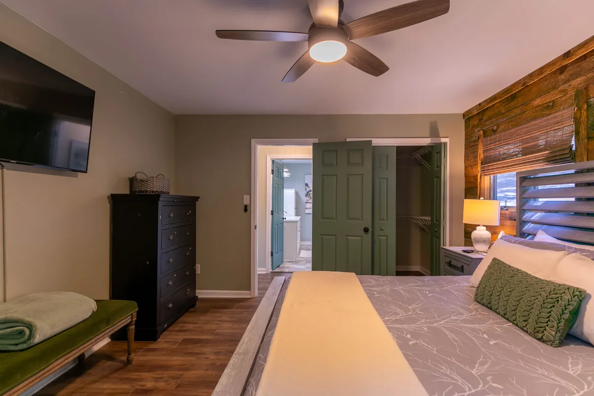 A bright and inviting bedroom with a comfortable bed dressed in decorative pillows, a green accent wall, and a wooden feature wall. The room includes a mounted television, perfect for relaxing with a movie before bed, and exudes a calming, restful mood.