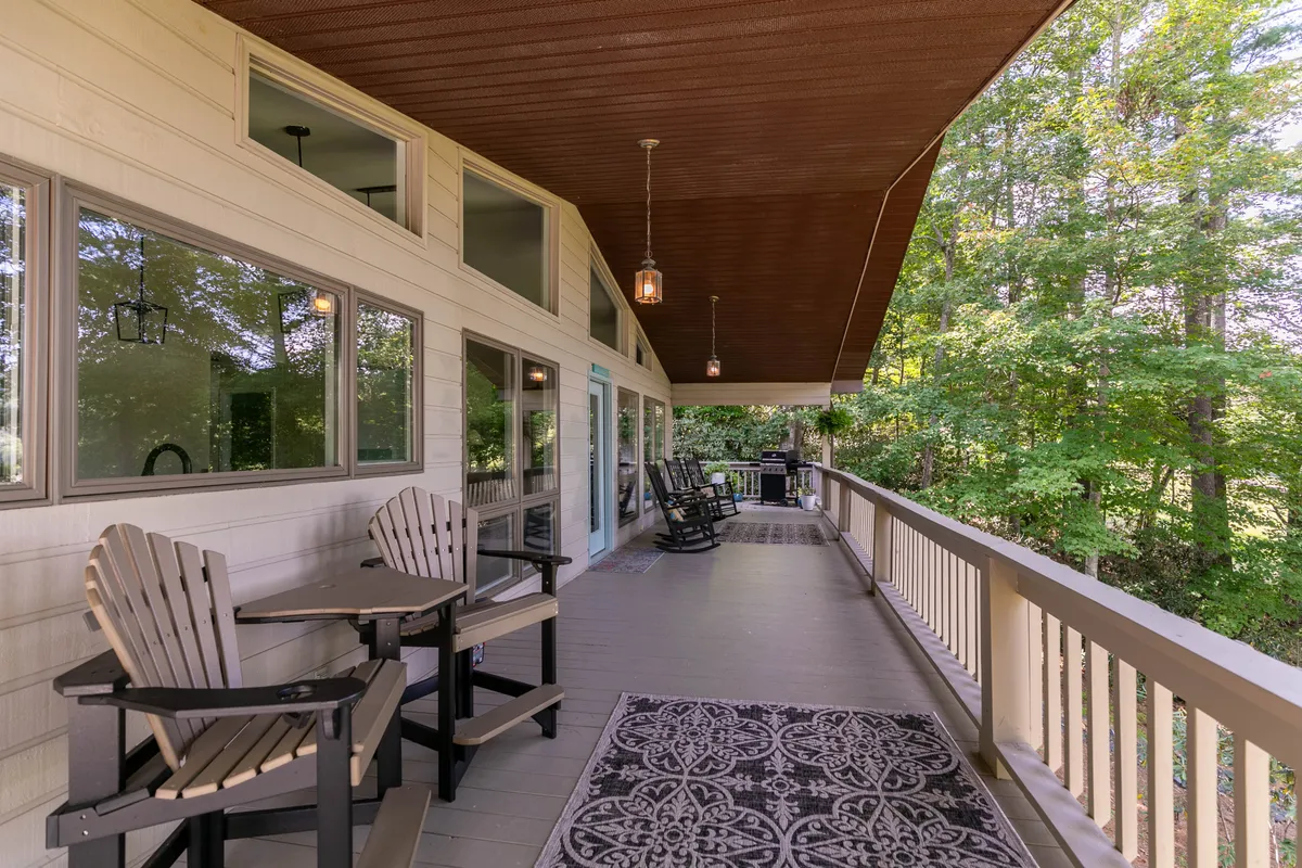 A serene outdoor dining area on the deck, with a large dining table surrounded by chairs, rocking chairs nearby, and hanging lanterns providing ambiance. Lush greenery and mountain views frame the space, making it perfect for al fresco meals under the stars or at sunrise.