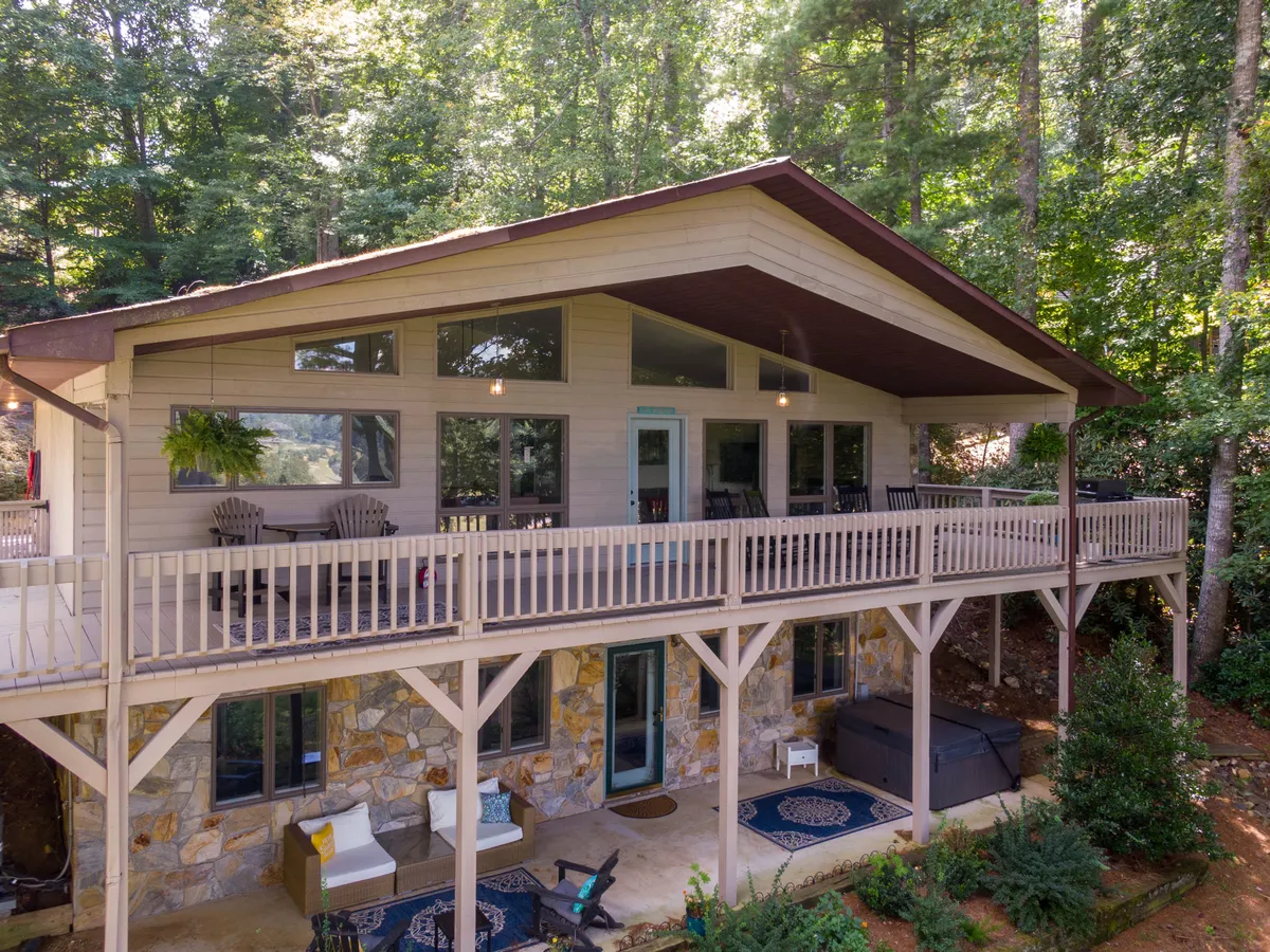 The image showcases the exterior of a charming cabin nestled among tall pines, featuring a spacious deck with comfortable seating, large windows that invite natural light, and beautiful natural stone accents. Lush greenery surrounds the property, creating a serene, private setting. The deck offers sweeping sunset views, perfect for relaxing, outdoor dining, and enjoying tranquil moments immersed in nature.