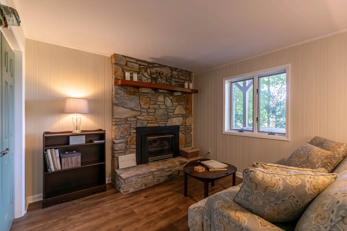 A cozy living space centered around a rustic stone fireplace, with a plush couch and a wooden shelf adorned with decorative items. A large window offers glimpses of the outside, evoking the warmth and comfort of cabin life during the chilly winter months.