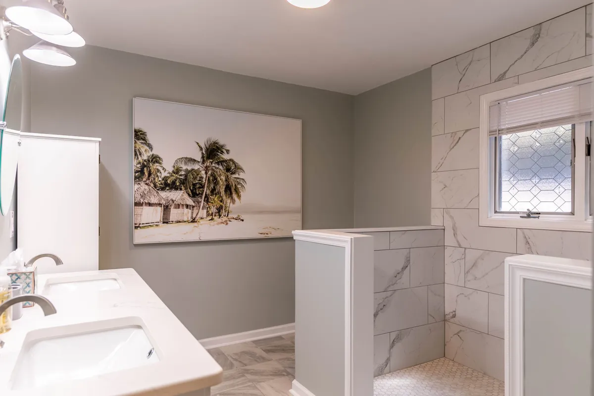 A modern, tranquil bathroom with double sinks, palm tree-themed artwork, and a walk-in shower. The space is flooded with natural light and offers ample room for morning routines or relaxing, spa-like showers.