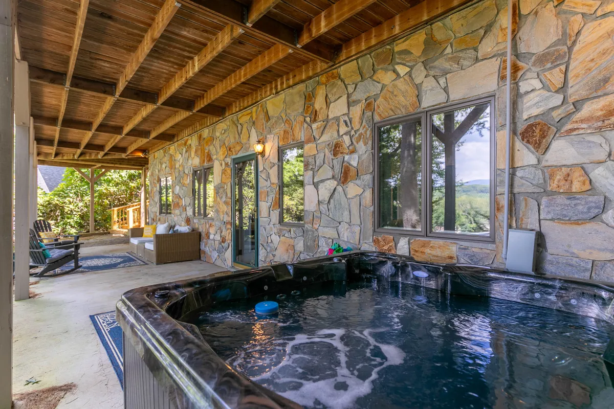 This image features an inviting hot tub set against a stone wall, with a cozy seating area nearby and large windows looking out onto the natural landscape. The scene highlights a tranquil environment perfect for relaxing and socializing outdoors, with glimpses of sunset lighting and the sense of being surrounded by nature.