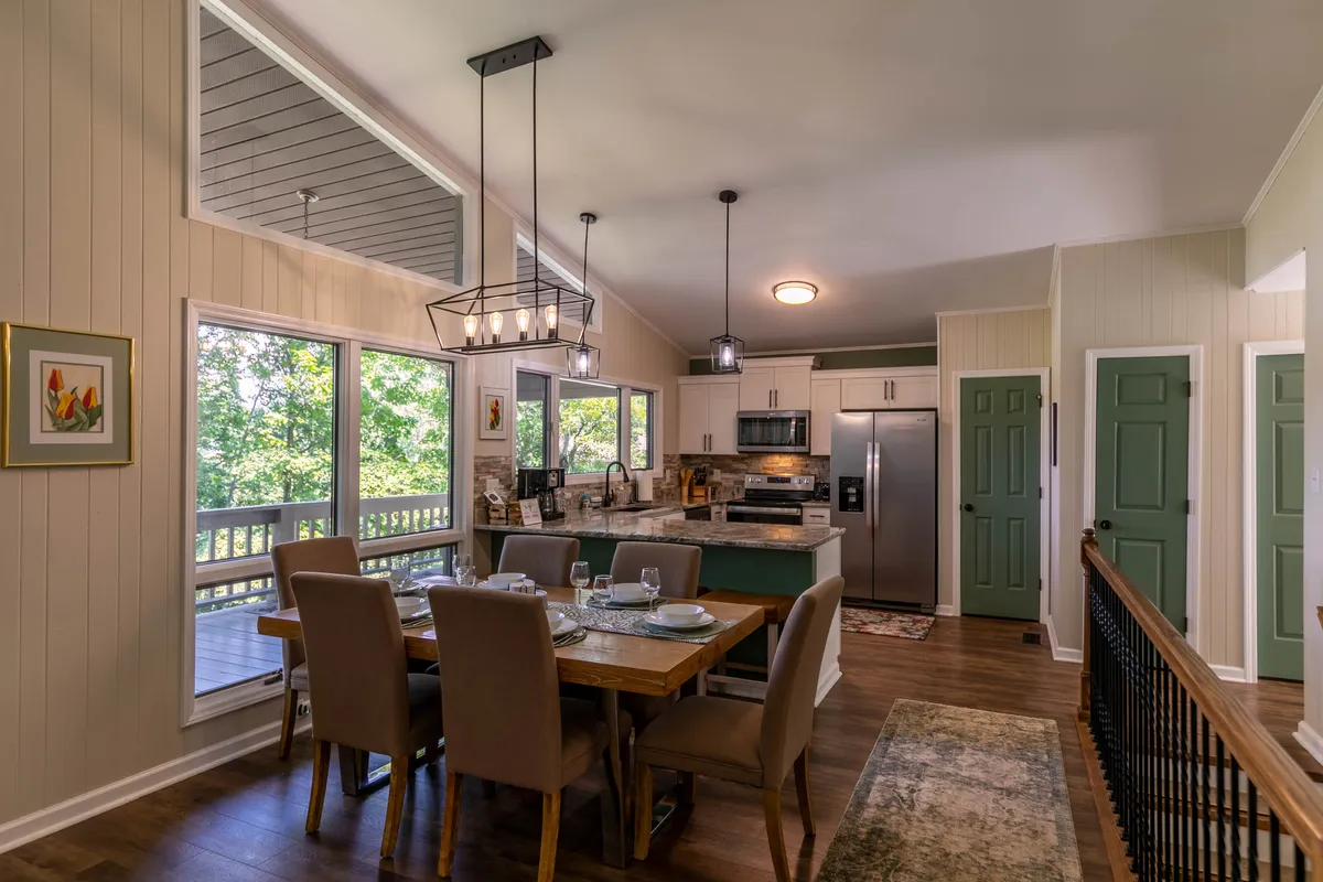 A bright, welcoming dining area seamlessly connected to a modern kitchen, featuring large windows that flood the space with natural light. The kitchen boasts stainless steel appliances, granite countertops, and a variety of cooking tools in view, while the dining table is set for a meal, inviting group gatherings.
