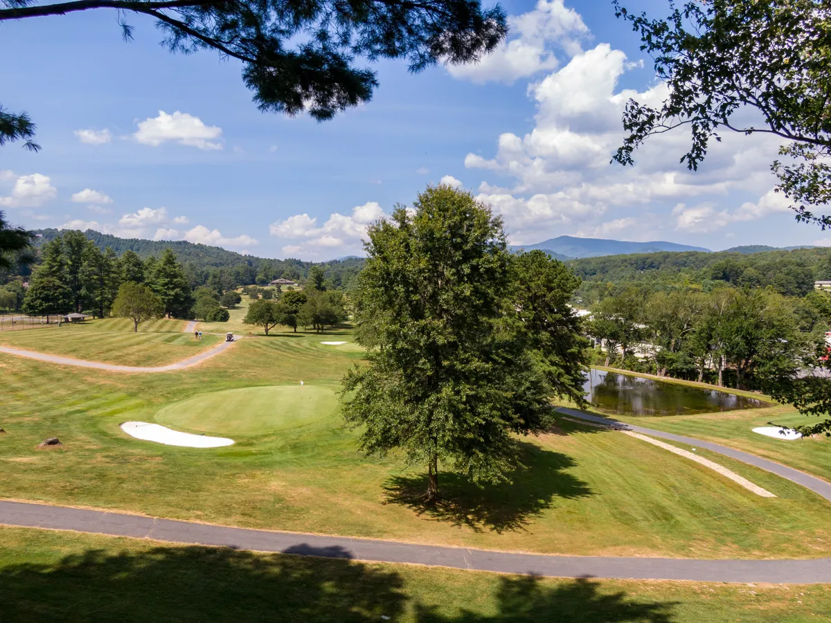 A sweeping vista of the golf course, with a tranquil pond, lush trees, and mountains rolling in the background. The scene blends the beauty of the course with the peacefulness of the surrounding nature, inviting guests to both play and relax.