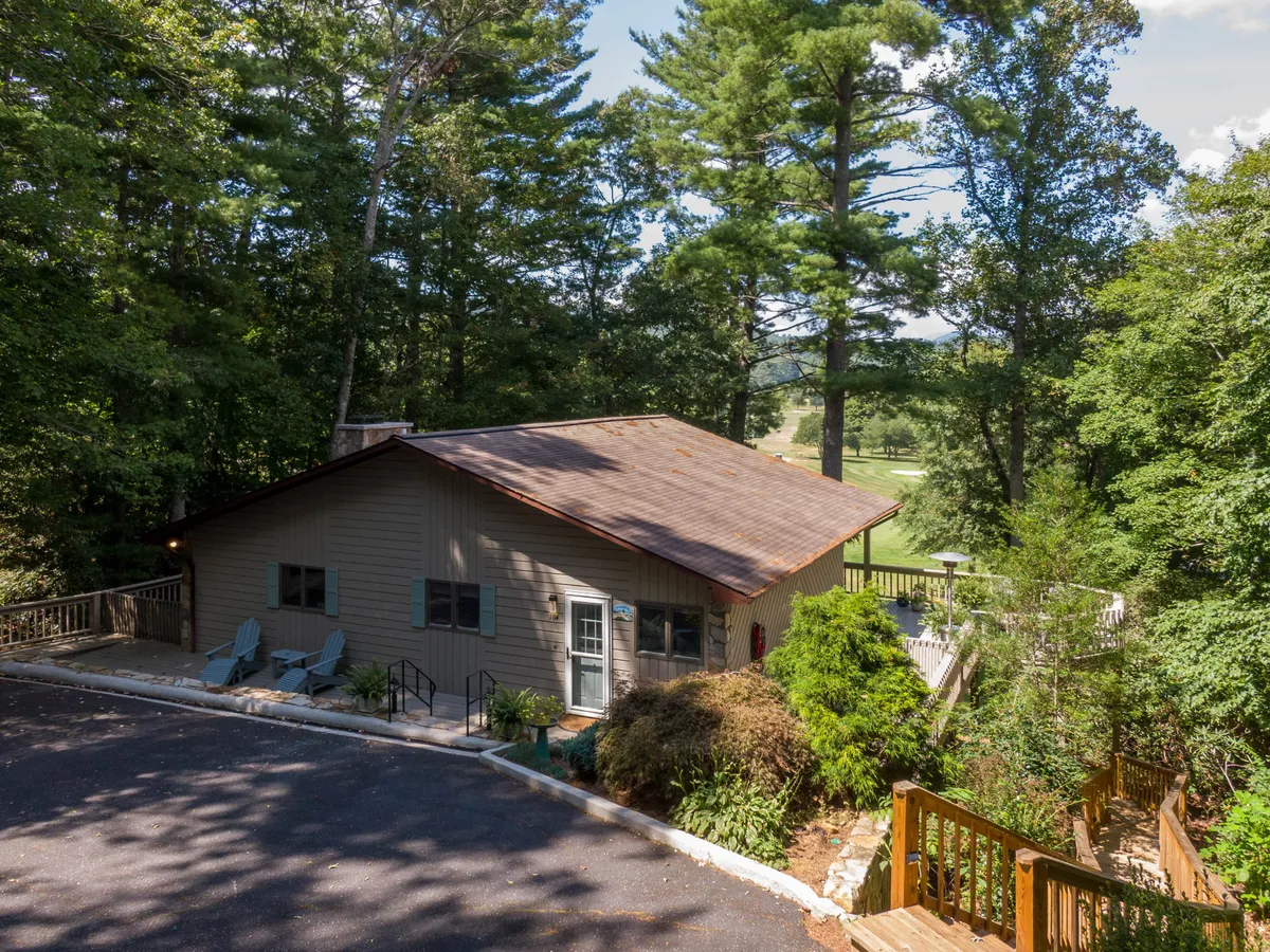 This exterior image displays a spacious house with a wrap-around deck nestled among trees. The deck is set for relaxation and overlooks a serene natural landscape, hinting at sunset views and ample space for family gatherings or quiet reflection. The peaceful mood and open design showcase a welcoming environment for outdoor dining and stargazing.
