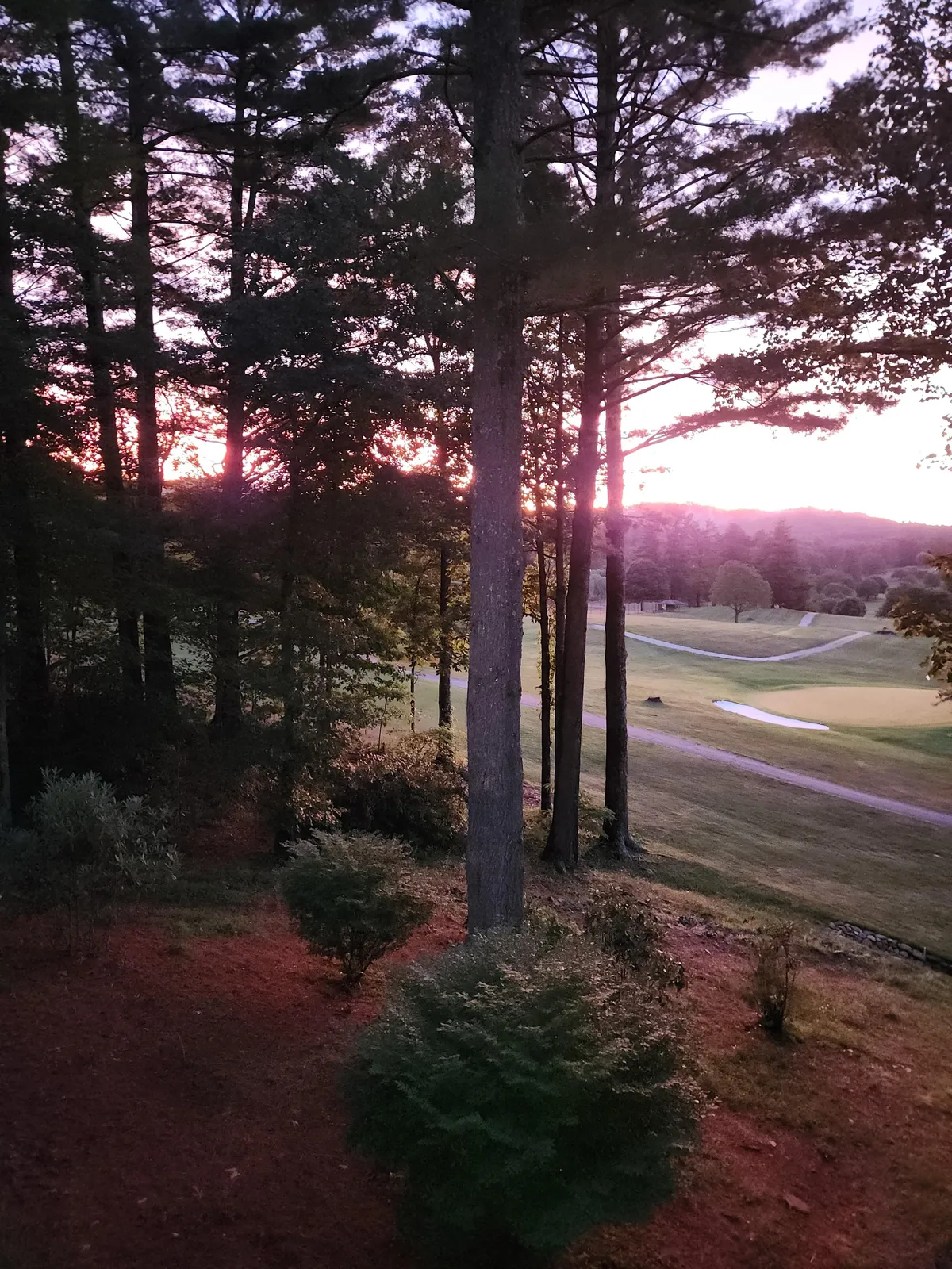 This image captures a scenic view of the golf course framed by tall trees and a vivid sunset sky. The fairway and greens are visible, making it clear that this property is situated directly alongside the golfing action, while the natural light sets a serene mood.